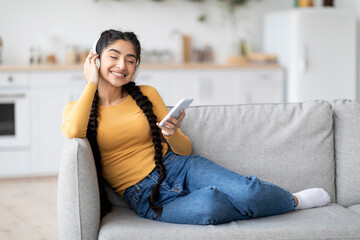 Obraz premium Happy Indian Female Wearing Wireless Headphones Relaxing With Smartphone At Home