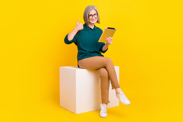 Full length photo of successful agent lady sit podium use tablet demonstrate thumb up isolated on yellow color background