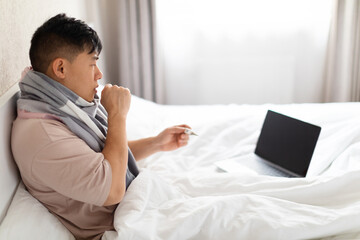 Asian man sitting in bed, coughing, using laptop, mockup