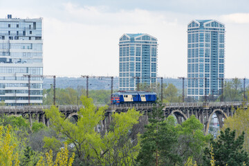Electric locomotive direction for modern city over arched bridge. Old train on the railway riding in Merefo-Kherson bridge across Dnieper river. Cityscape of new high-rise buildings and blue carriage.