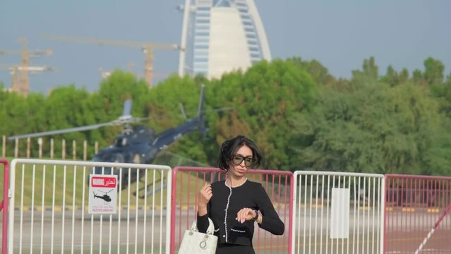 A young woman in a formal black dress walks through the airfield against the background of a small private helicopter flying by.