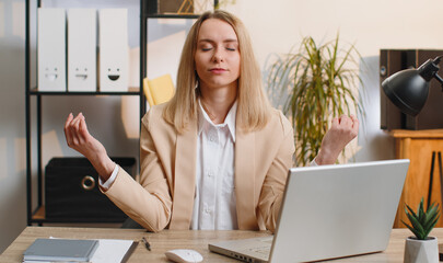 Young businesswoman working on laptop computer, meditating, doing yoga breathing exercise in lotus...