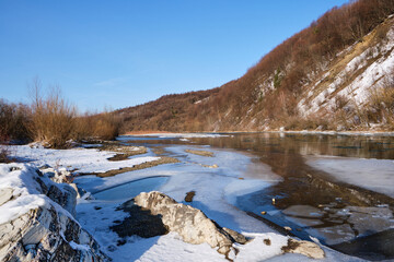 Winter landscape on a clear day. A view of a mountain river with ice and mountains covered with snow and stones with different structures and a clear sky