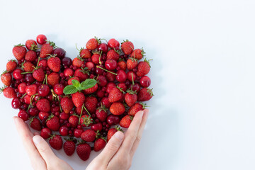 Female hands holding fresh strawberries and cherries arranged in heart shape on white background. Top view, copy space. Spring and summer fruit harvest, love for plant-based food concept.
