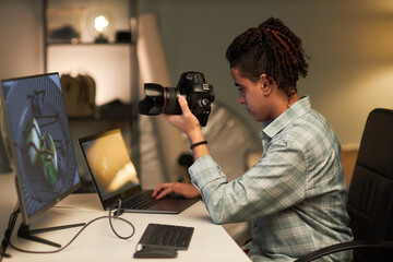 Young photographer with camera sitting at his workplace in front of computer monitor and using laptop