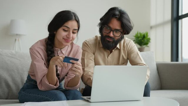 Arabian Indian muslim couple family sit on sofa in living room home office use credit bank card typing code number in laptop banking app pay online multiethnic woman and man shopping internet delivery