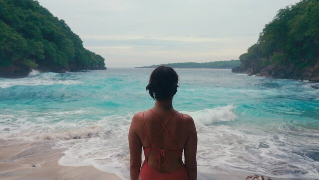 A lonely woman with short hair standing on wild ocean beach with turquoise water looking on waves. Pensive thoughtful person on seaside. Relaxation and enjoying nature on summer holidays. Middle plan