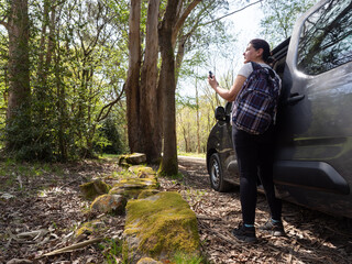 middle-aged woman alone with backpack, gps in hand looking at the trail beside a van with the door open on a road in the fore