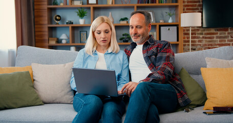 Happy middle aged couple looking at laptop screen sitting on sofa at home. Woman and man in living room searching internet typing online using computer watching video surfing internet.