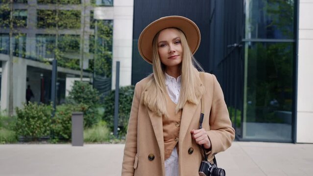 Portrait positive stylish female photographer standing outdoors in modern city street. Beautiful caucasian blonde woman wearing beige coat and hat posing with retro style photo camera on her shoulder