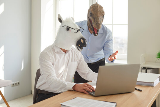 Funny animal people work in office. Two men in unusual disguise use laptop. Man wearing horse mask and glasses types on notebook computer while his silly dinosaur coworker tells him what to do