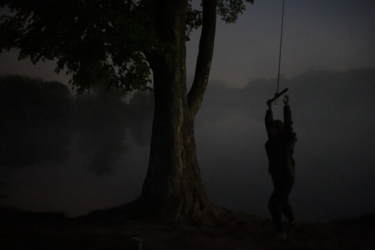 Man holding a swing at late foggy night next to a ancient tree 