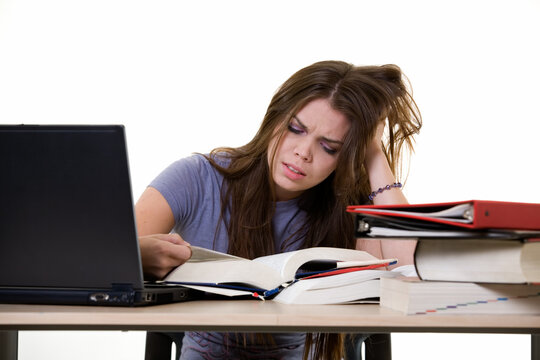 Young woman sitting in front of laptop beside a pile of thick textbooks while reading one with a frustrated stressed expression
