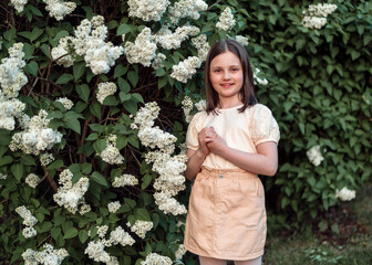 Portrait of a girl near flowering white lilac trees. A beautiful girl of 8 years old in beige clothes and with a bob haircut at sunset in the spring.