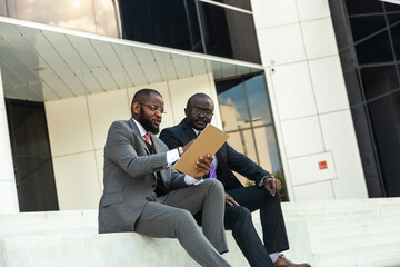 Friendly meeting of business partners outdoors. Two dark-skinned men in suits sit on the steps of a city building with a notebook and have a conversation. Working break. Support and collaboration