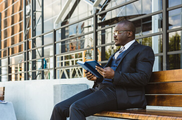 A dark-skinned businessman in a suit reads an outdoor notebook while sitting on a bench against the backdrop of city buildings