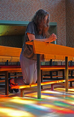 A woman kneeling in prayer in a church. Sunlight streams through the stained glass windows and...