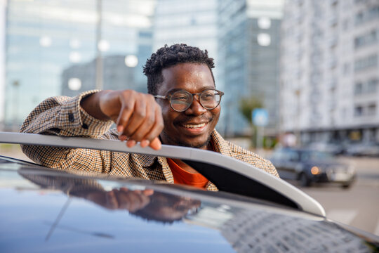Smiling man in glasses leaning on car roof in city
