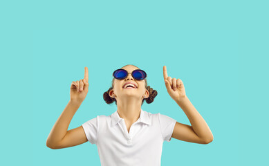 Cheerful little Caucasian girl teenager in funny sunglasses looks up in amazement pointing fingers...