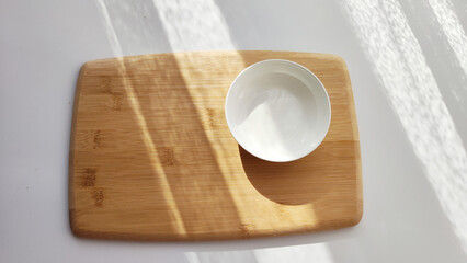 Cutting board made of natural bamboo and white plate saucer on table with shade and light from sun. Abstract natural background, texture, frame, copy space. Concept of cooking with beautiful things