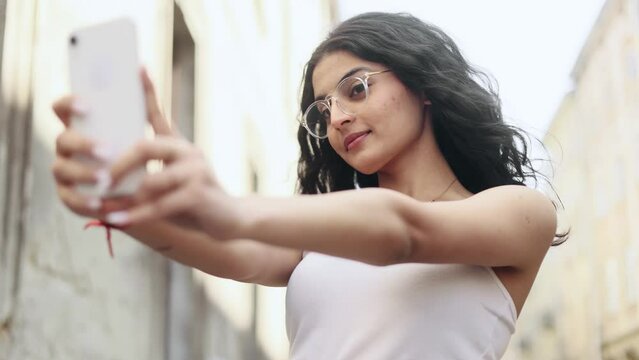 Portrait of young indian curly woman grimacing taking selfie photograph saving great memories on smartphone mobile phone at city centre Pretty girl in glasses create content for social media outdoors