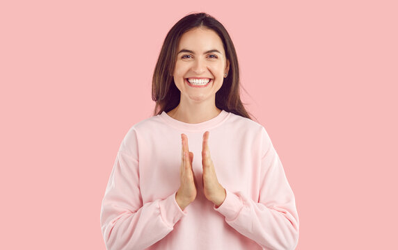 Studio portrait of happy applauding young brunette woman. Joyful girl with toothy smile wearing light pink sweatshirt clapping her hands on isolated studio background looking at camera
