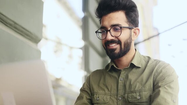 Handsome young indian businessman freelancer with glasses working on laptop and looking at camera at coffee shop Busy focused man have distance remote freelance work on computer outdoors alone