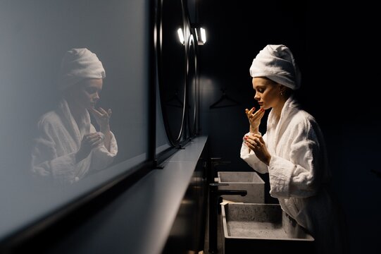 young Woman in bathrobe applying cream on face in bathroom
