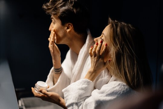 Lady and boyfriend applying cream on faces near mirror 
