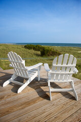 Two Adirondack style chairs sitting on a wooden deck, facing the shore. The grassy beach is in view beyond the deck. Vertical shot.