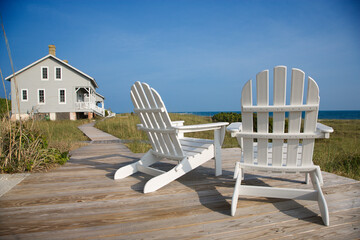 Two Adirondack style chairs sitting on a wooden deck, facing the shore. There is a large home in...