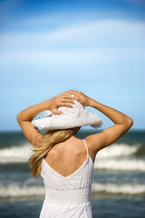 Young woman holds her hat down as she looks out at the ocean from the beach. Vertical shot.