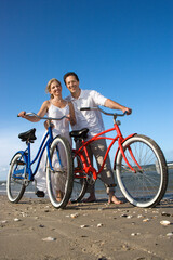 Smiling man and woman pose with bicycles on the sand near the beach. Vertical shot.