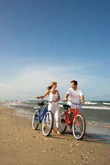 Fototapeta premium Smiling man and woman walk bicycles down the beach coast. Vertical shot.