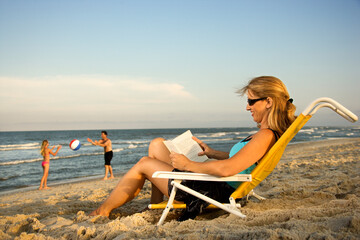 Woman at the beach reads a book while her family plays in the background by the ocean.