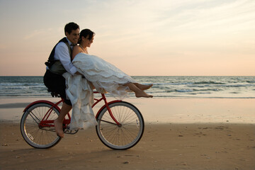 Bride rides the handle bars of a bicycle being driven by her groom on beach. Horizontal shot. - Powered by Adobe