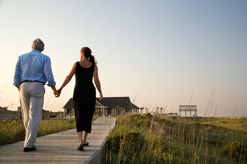 Couple walk hand in hand on a boardwalk towards a beach pavilion. Horizontal shot.