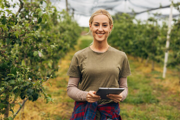 A Caucasian blond woman is standing in the orchard and holding a tablet.