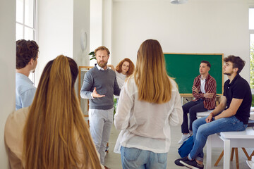 Naklejka na ściany i meble Smart and intelligent male teacher speaks in classroom surrounded by his high school students. Man talks to students at break or informal meeting where they discuss ideas about educational process.