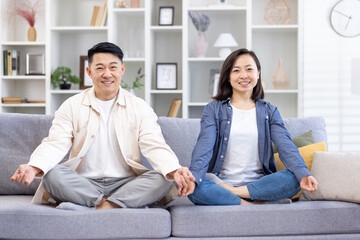 Portrait of a young Asian family, a couple doing yoga at home. A man and a woman are sitting on a sofa in the lotus position and smiling at the camera. take care of their mental health.