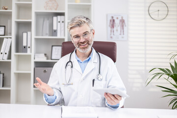 Portrait of a smiling senior male doctor who remotely consults patients online, sits in the office at the table and talks to the camera on a video call