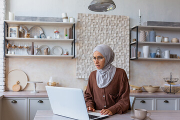 Remote work. Muslim young woman in hijab freelancer working from home online on laptop. Sitting in the kitchen at the table and looking worriedly and confusedly at the monitor.