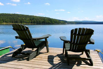 Two adirondack wooden chairs on dock facing a blue lake with clouds reflections