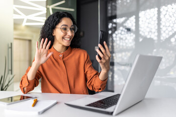 Young Muslim woman sitting in the office at the table, working on the laptop and talking on the phone on a video call, smiling, greeting, waving at the camera.