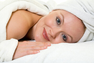 Fototapeta na wymiar Attractive woman laying on stomach on a white pillow wearing white robe and towel on head with a bare shoulder and happy expression