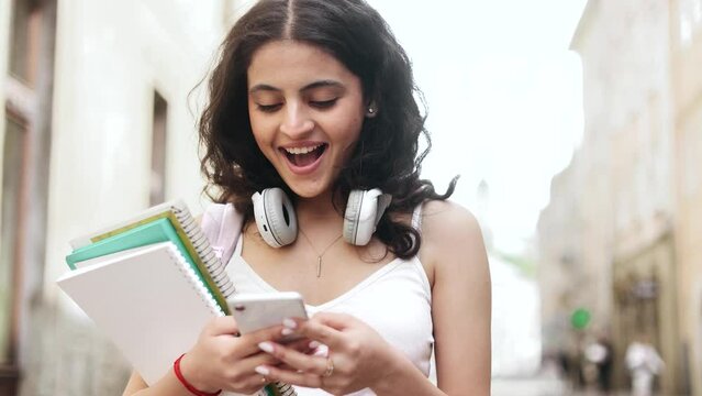Amazed young indian woman student extremely happy after read message on smartphone at city street after lessons Excited girl looking at phone screen and pleased rejoices received great news outdoors