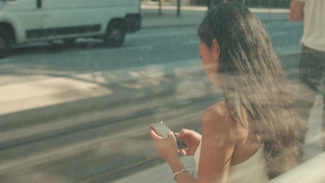 Young woman sits at public transport stop, uses mobile phone, blurry view through the glass