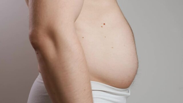 A man with a bare torso in white underpants inflates and sticks out his big belly. The concept of weight gain, overeating and obesity. Side view