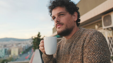 Peaceful guy looks at the city from the balcony and drinks coffee, enjoys the beautiful view