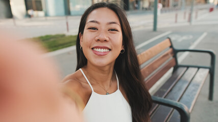 Pretty brunette girl dressed in white top and jeans is smiling making selfie while sitting on bench on cityscape background